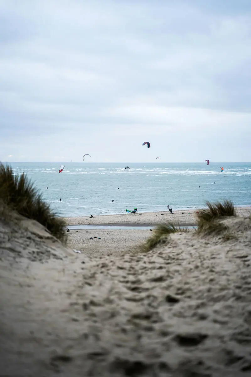 Dünenpfad zum Strand in Dänemark mit Kitesurfern über dem Meer und bewölktem Himmel.