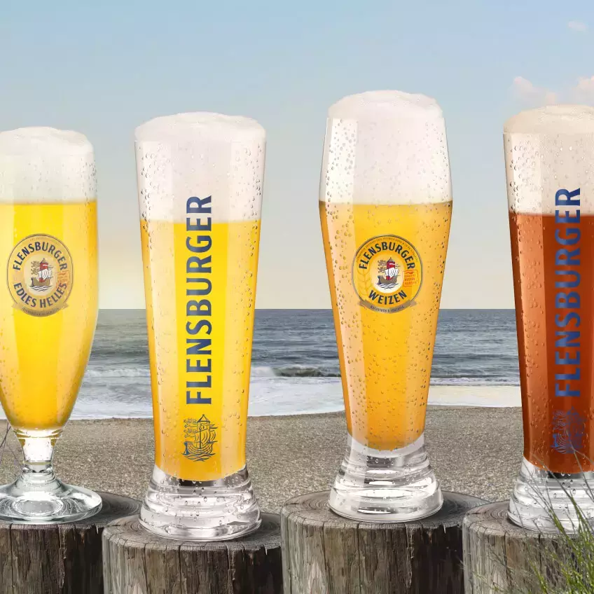 Vier Gläser mit unterschiedlich hellen Bieren auf Pollern am Strand.