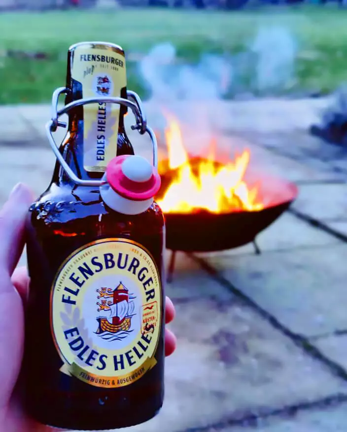 Eine Hand hält eine Flasche Flensburger Edles Helles, im Hintergrund eine Feuerschale mit Feuer.