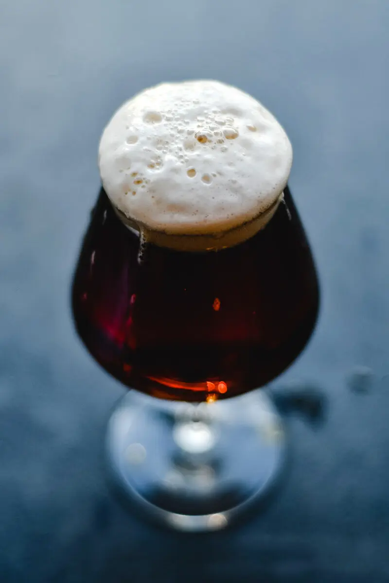 Nahaufnahme von dunklem Bier im Tulpenglas mit cremiger Schaumkrone auf blauem Hintergrund.