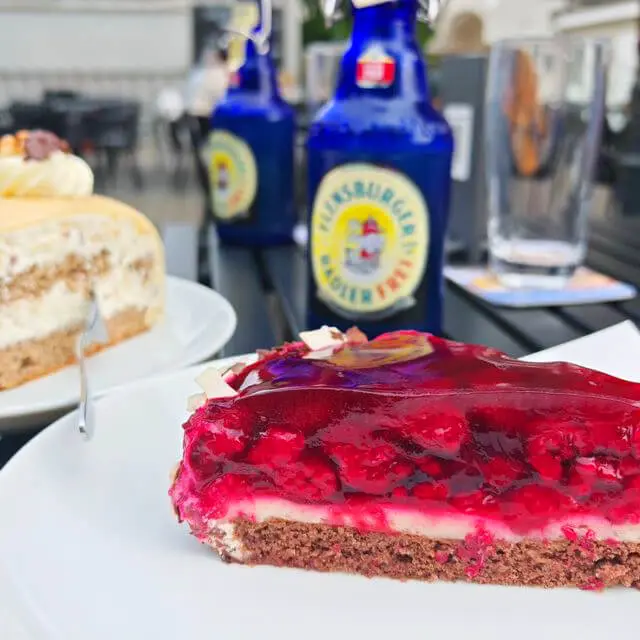 Stück Himbeertorte mit Gelee und Sahne auf Teigboden, Teller vor blauen Radler-Frei-Flaschen von FLENS.