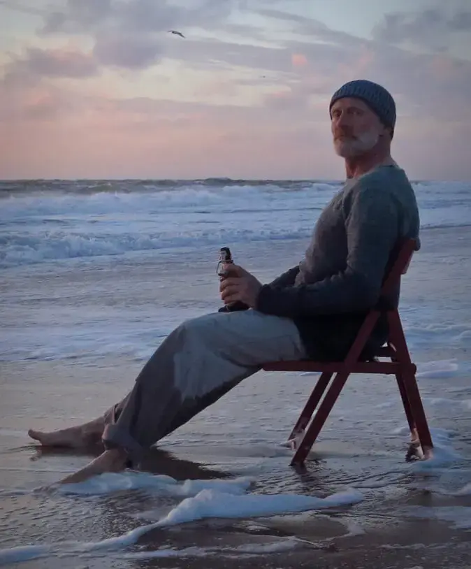 Andreas Arndt sitzt auf Klappstuhl am Meer bei Sonnenuntergang, hält die Beine im flachen Wasser und hält FLENS Flasche in der Hand.