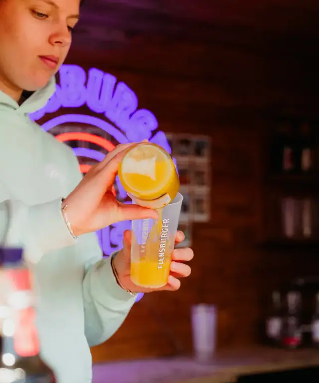 Person gießt Flensburger Küstenlimo Orange in einen Flensburger-Becher am Tresen mit neonbeleuchtetem Küstenlimo-Logo.