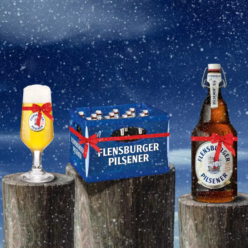 Bierglas, Mega-Plops von FLENS und eine Kiste Bier auf Holzpollern im Schnee und mit Geschenkeband.