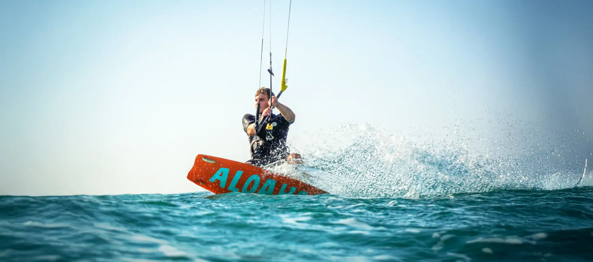 Kitesurfer auf Aloahoi-Surfboard fährt durch Wellen, große Wasserspritzer im türkisblauen Meer.