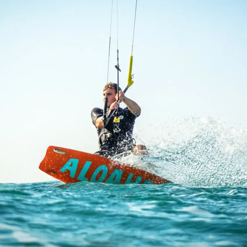Kitesurfer auf Aloahoi-Surfboard fährt durch Wellen, große Wasserspritzer im türkisblauen Meer.