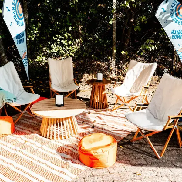 Gemütliche Outdoor-Lounge mit Klappstühlen, Holz-Tisch, orangenen Sitzkissen und gestreiftem Teppich.