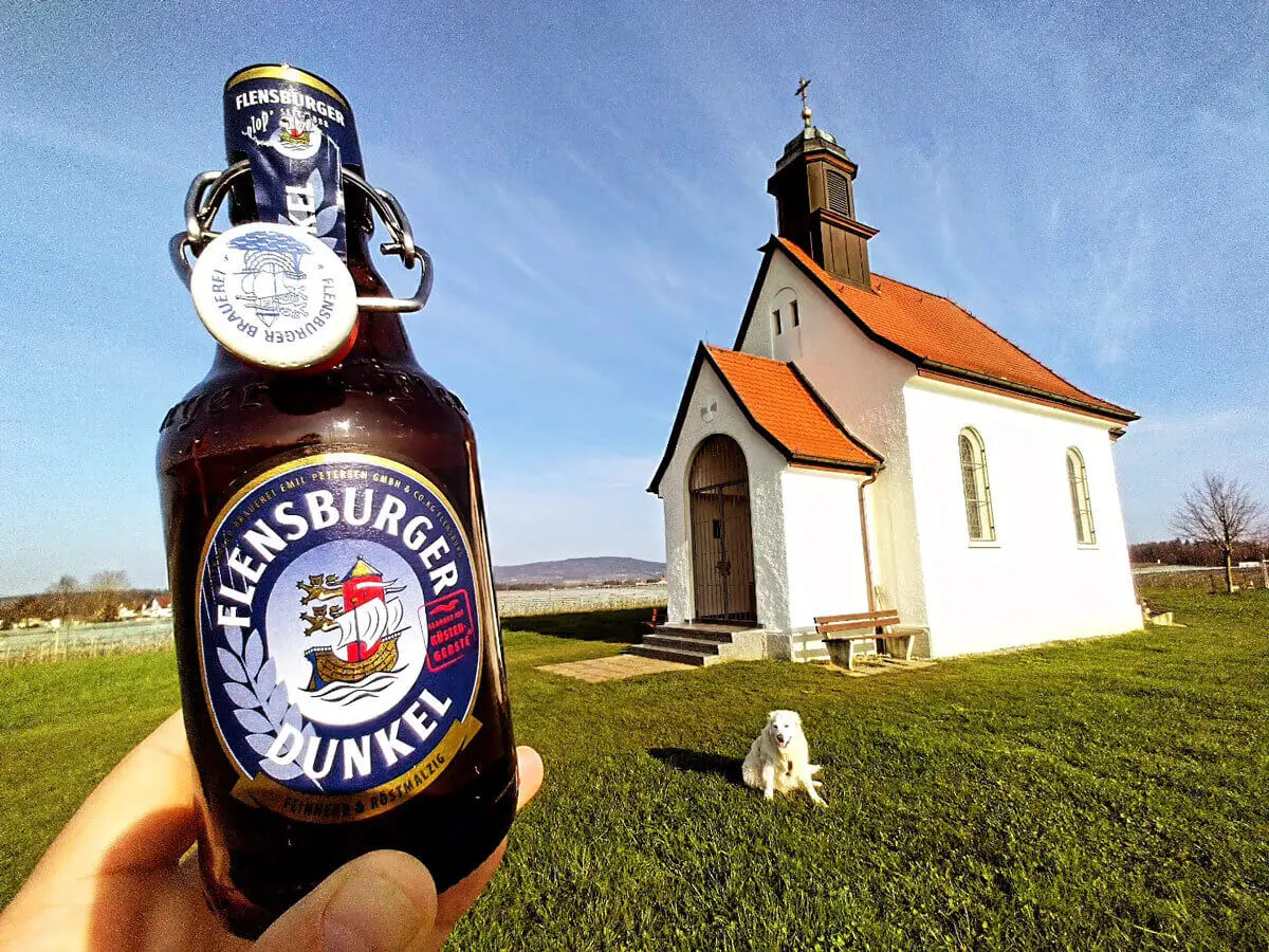 Flensburger-Dunkel-Bierflasche im Vordergrund vor weißer Kapelle mit Hund auf grüner Wiese.