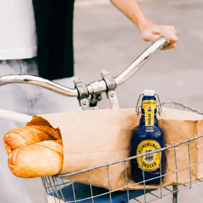 Fahrradkorb mit Papiertüte, frischem Baguette und blauer FLENS Radler-Flasche.