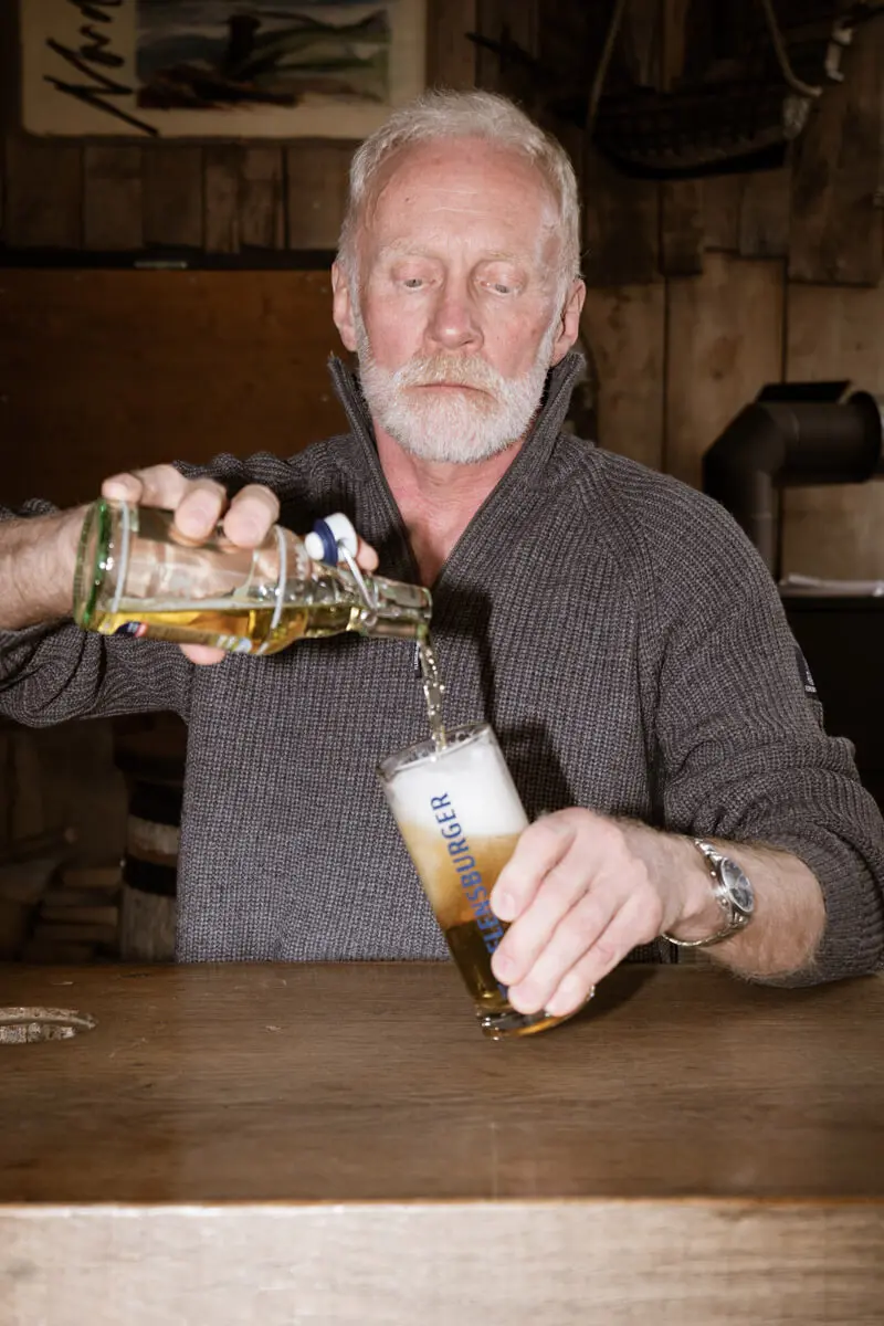 Andreas Arndt in grauem Strickpullover beim Bier-Einschenken in Glas an rustikalem Holztresen.