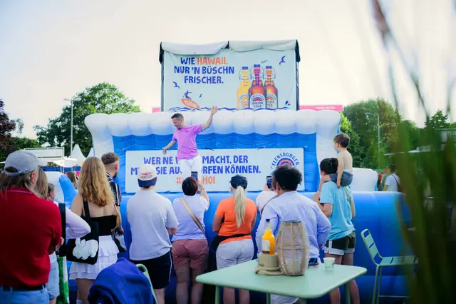 Kieler Woche 2025 mit aufblasbarem Bullriding-Element mit Surfbrett: Kind surft, Publikum schaut zu, großes Banner der Küstenlimos im Hintergrund.