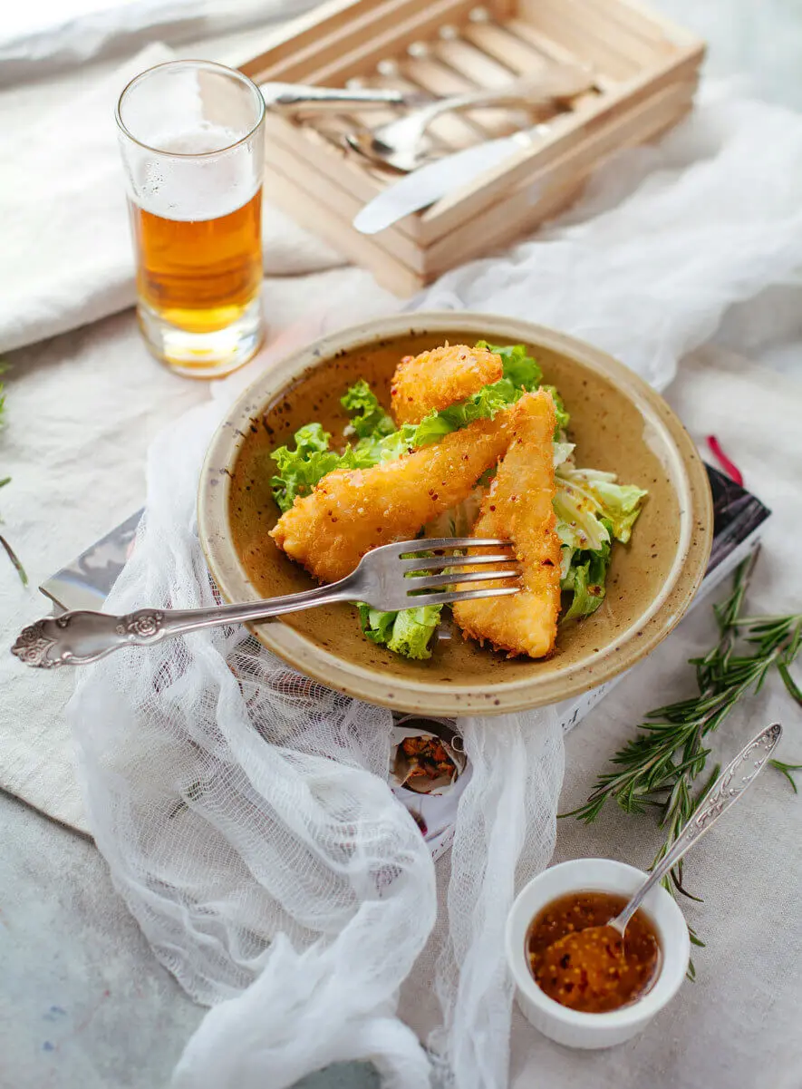 Knusprige Backfischdreiecke mit heller Panade auf Blattsalat mit Chili-Dip und Bierglas.