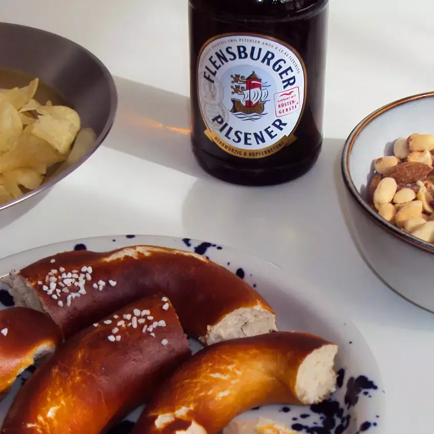 Drei Schalen mit Snacks wie Brezeln, Nüssen und Chips, in der Mitte eine Flasche Flensburger Pilsener.