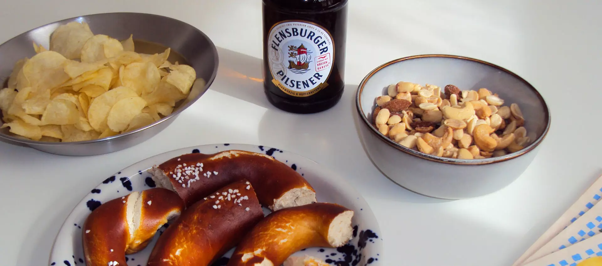 Drei Schalen mit Snacks wie Brezeln, Nüssen und Chips, in der Mitte eine Flasche Flensburger Pilsener.