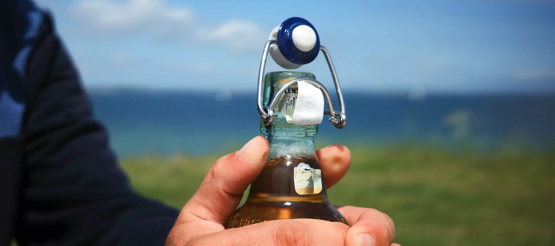 Close-up vom Flensburger Gold, wie die Flasche geöffnet wird, dahinter verschwommen eine Küstenlandschaft.
