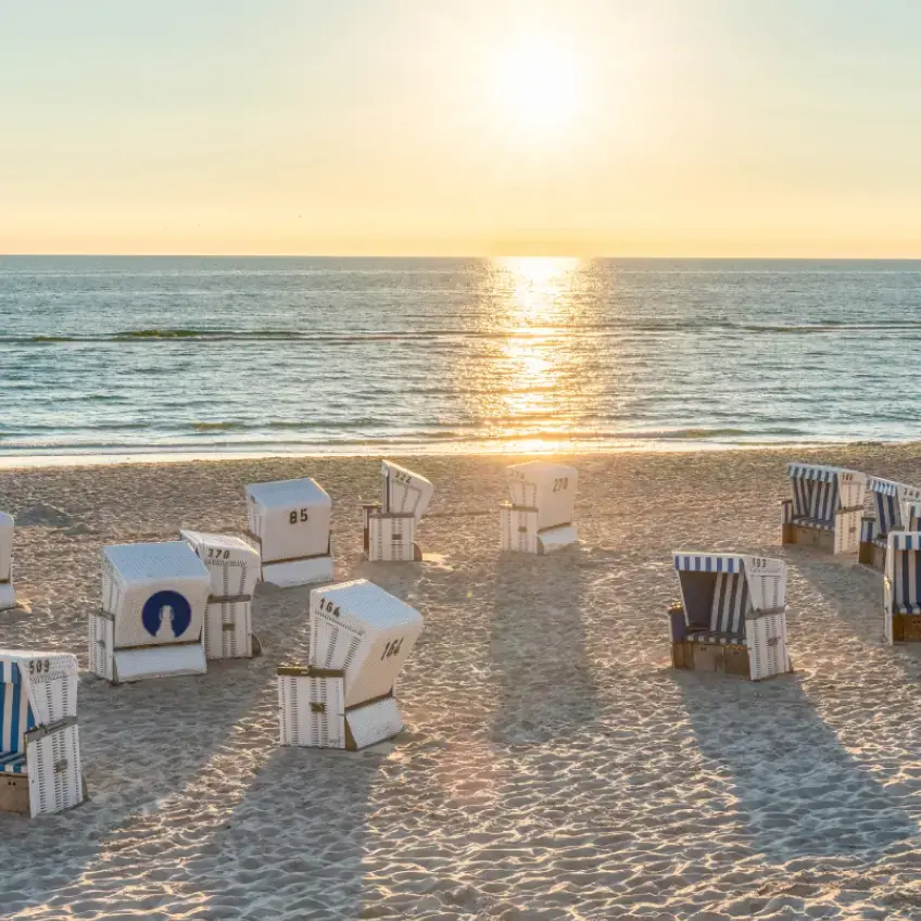 Weiß-blaue FLENS Strandkörbe auf Sandstrand bei Sonnenuntergang, ruhiges Meer mit Sonnenreflexion.