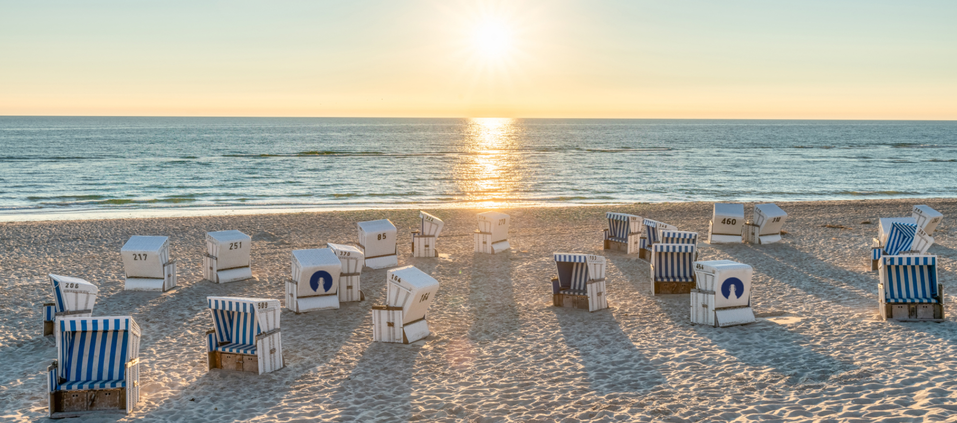 Weiß-blaue FLENS Strandkörbe auf Sandstrand bei Sonnenuntergang, ruhiges Meer mit Sonnenreflexion.