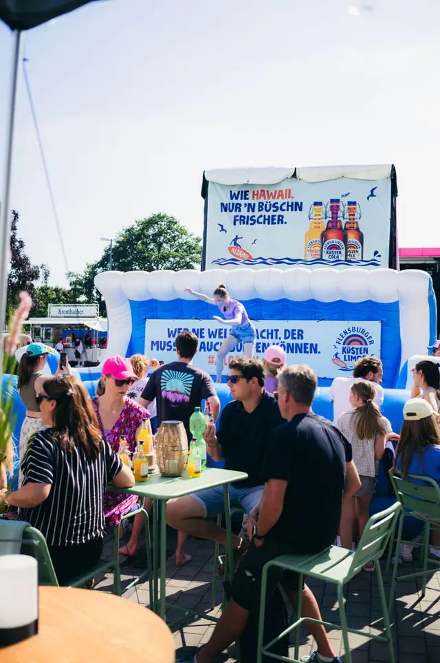 Kieler Woche 2025 mit aufblasbarem Bullriding-Element mit Surfbrett, Frau surft auf dem Surfbrett, davor Menschen mit Küstenlimo-Getränken an den Tischen.
