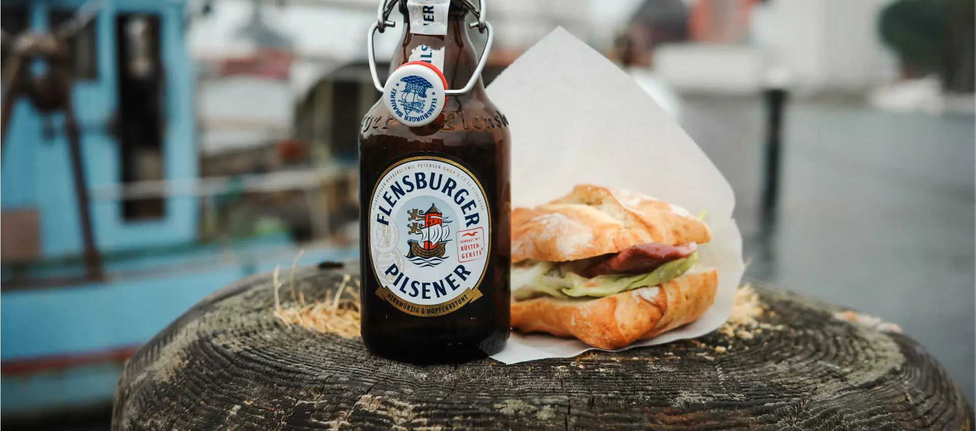 Flasche Flensburger Pilsener und ein Fischbrötchen auf einem Holzsockel im Hafen von Flensburg.