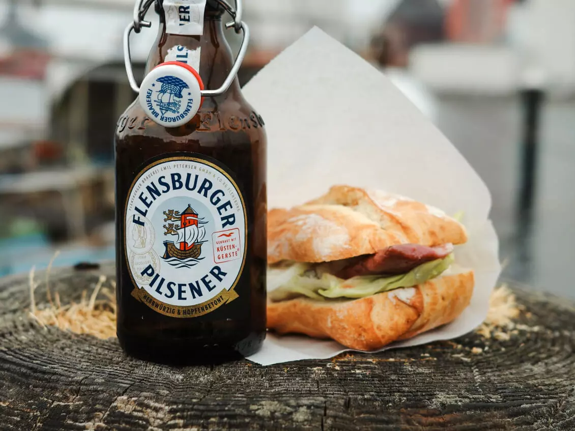 Flasche Flensburger Pilsener und ein Fischbrötchen auf einem Holzsockel im Hafen von Flensburg.