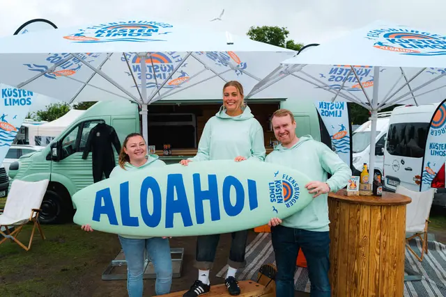Drei Menschen halten ein türkisfarbenes Surfbrett mit blauem Aloahoi-Schriftzug vor dem Promo-Van der Küstenlimos.