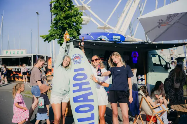 Gruppe junger Menschen feiert auf der Kieler Woche 2025 mit Küstenlimos, Surfbrett-Deko und Riesenrad im Hintergrund.