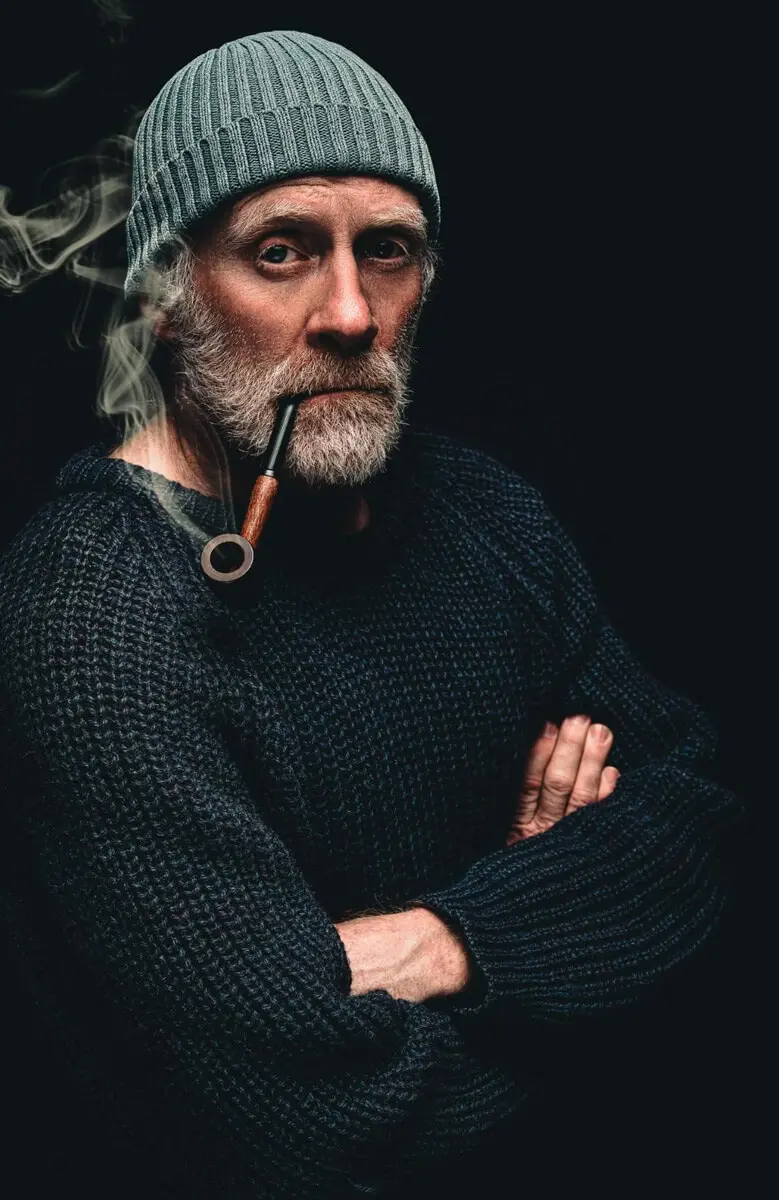 Andreas Arndt mit Strickmütze und in dunklem Pullover, raucht Pfeife vor dunklem Hintergrund.