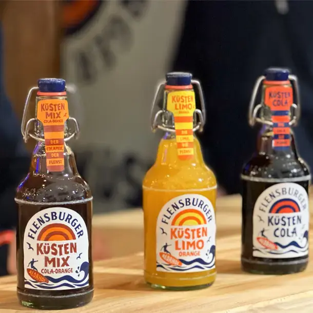Die drei Küstenlimoprodukte auf einem Tresen aus Holz: Flensburger Küstenmix, Küstenlimo Orange und Küstencola.