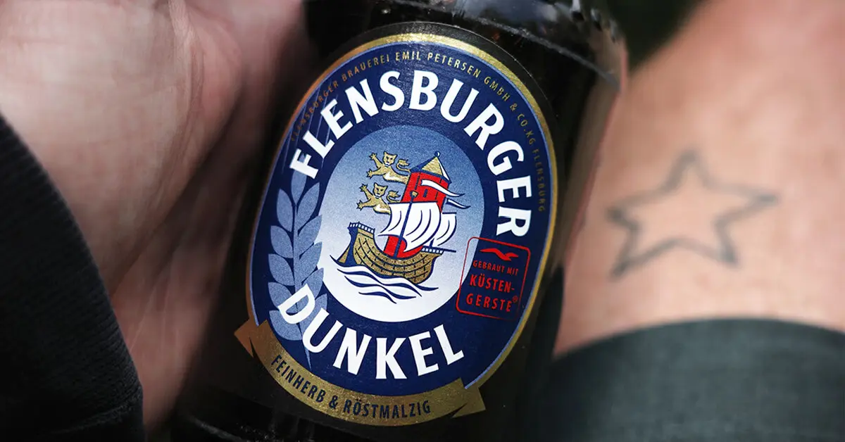Nahaufnahme, wie jemand eine Flensburger-Dunkel-Bierflasche in der Hand hält, im Hintergrund ein Oberschenkel mit einem Stern-Tattoo.