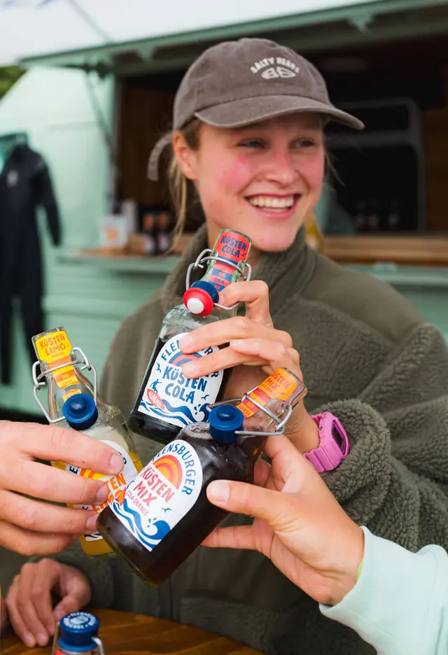Eine junge Frau stößt mit zwei anderen mit den Flensburger Küstenlimos an.