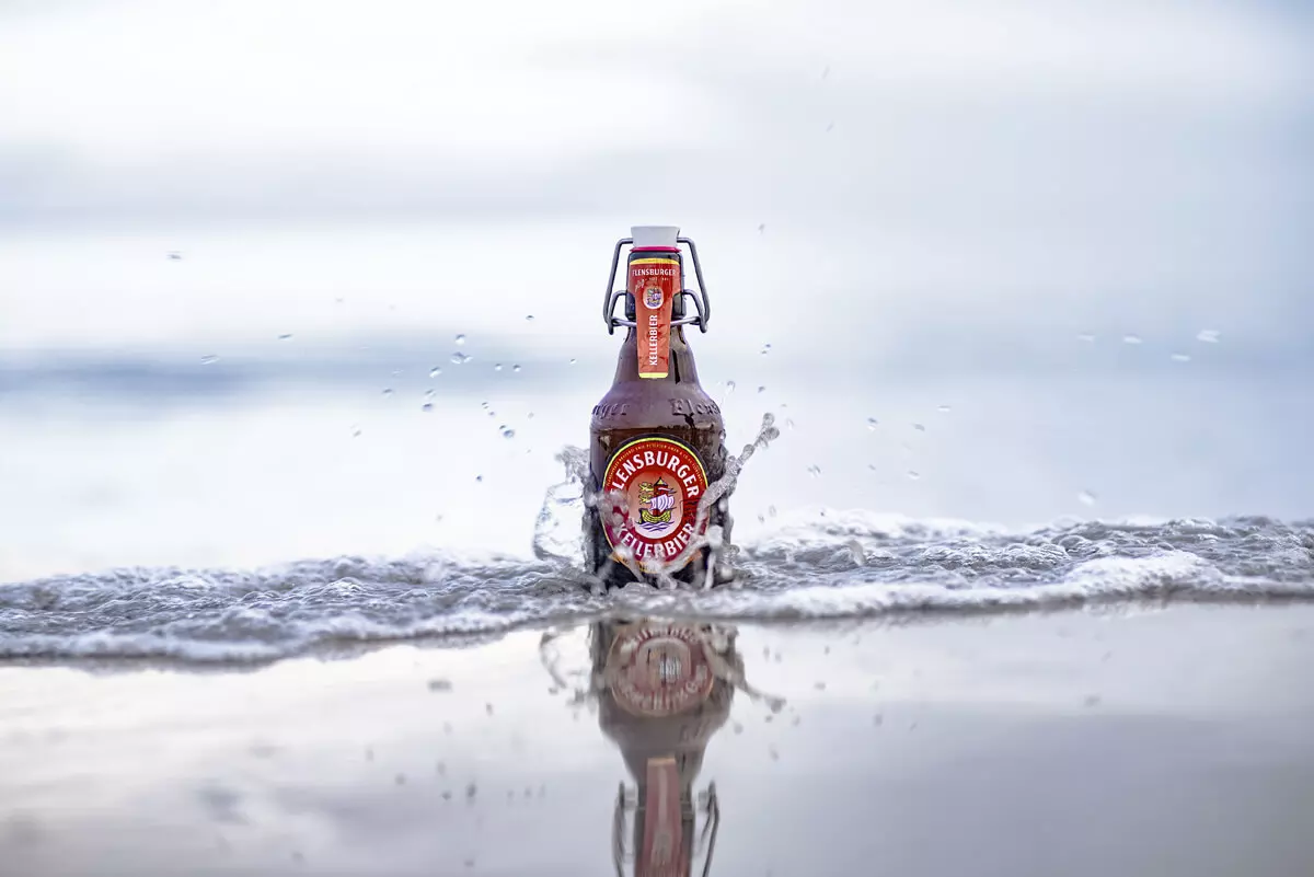 Eine einzelne Flasche Flensburger Kellerbier in der Welle am Strand.