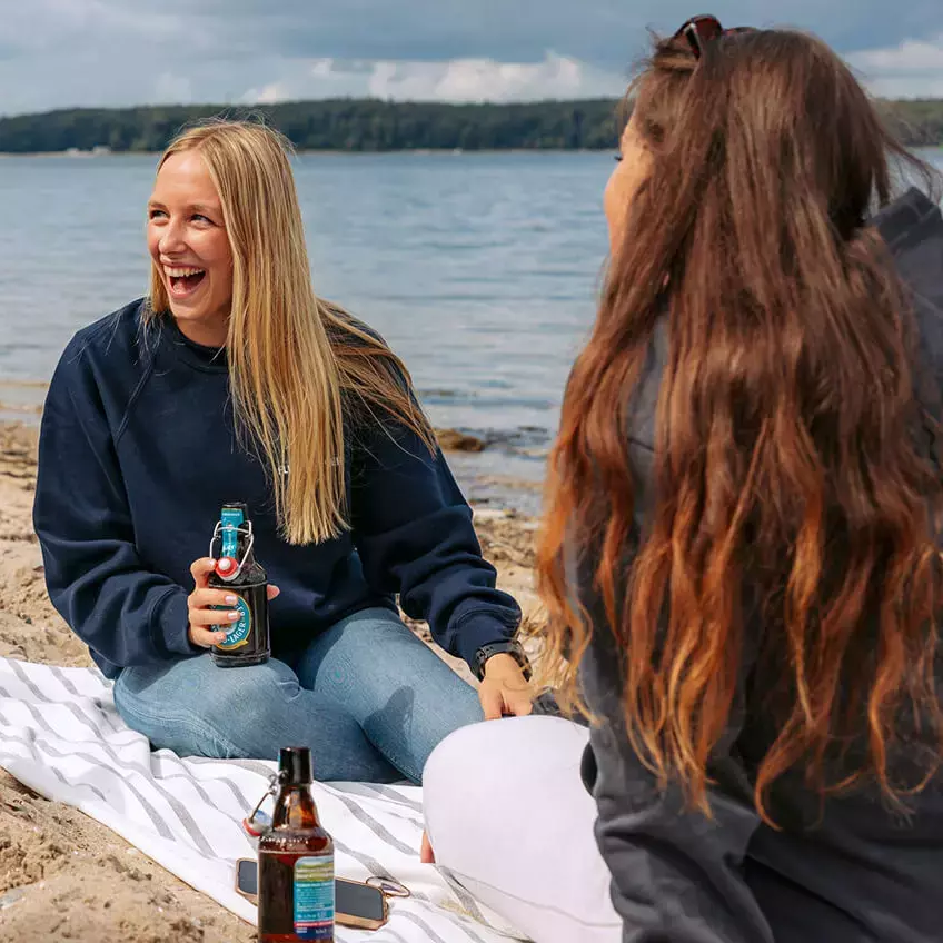 Eine Gruppe junger Frauen sitzt am Strand, hat eine gute Zeit und trinkt FLENS.