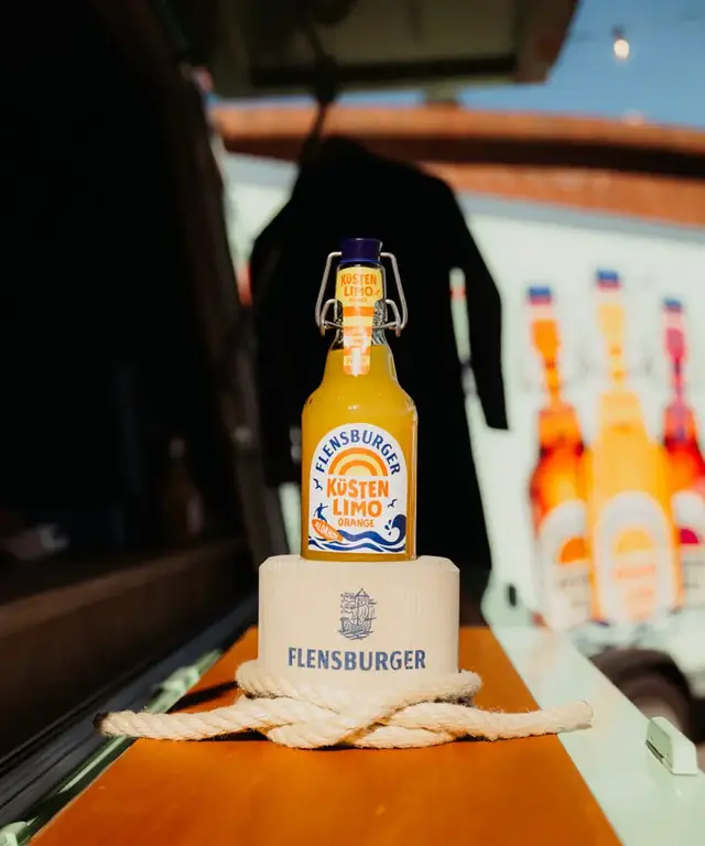 Flensburger Küstenlimo Orange auf Holzpodest mit Seildeko, im Hintergrund ein Neoprenanzug.