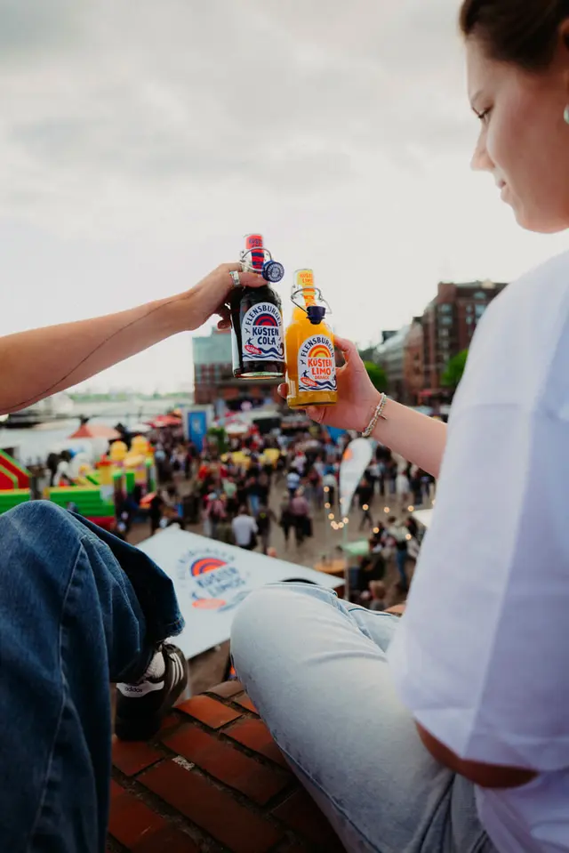 Zwei junge Menschen stoßen mit Flensburger Küsten-Cola und Küstenlimo Orange am Hamburger Fischmarkt und mit Menschenmenge im Hintergrund an.