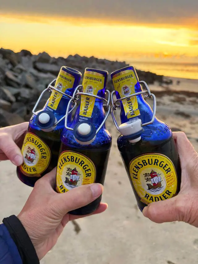 Drei Flensburger-Radler-Bügelflaschen beim Anstoßen am Strand bei Sonnenuntergang.