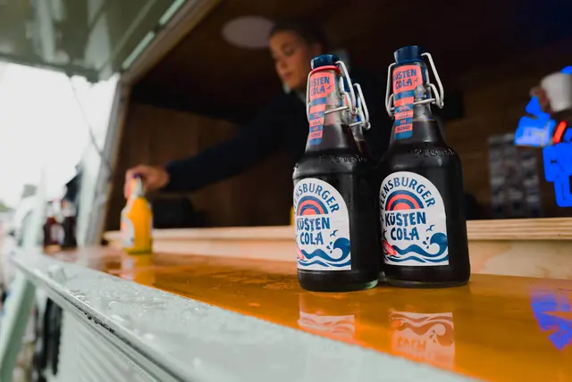 Zwei Küstencola-Flaschen mit Bügelverschluss auf nasser Holztheke des Küstenlimo-Vans.
