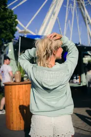 Junge Frau in mintfarbenem Hoodie mit ALOAHOI-Aufdruck, Rückenansicht, Kieler Woche und Riesenrad im Hintergrund.