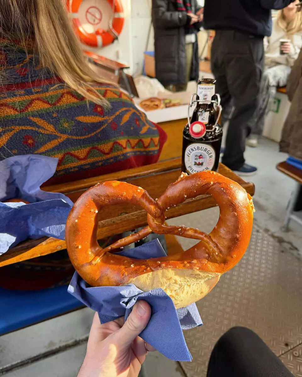 Eine dicke Brezel wird ins Bild gehalten, dahinter eine Flasche Flensburger Pilsener.