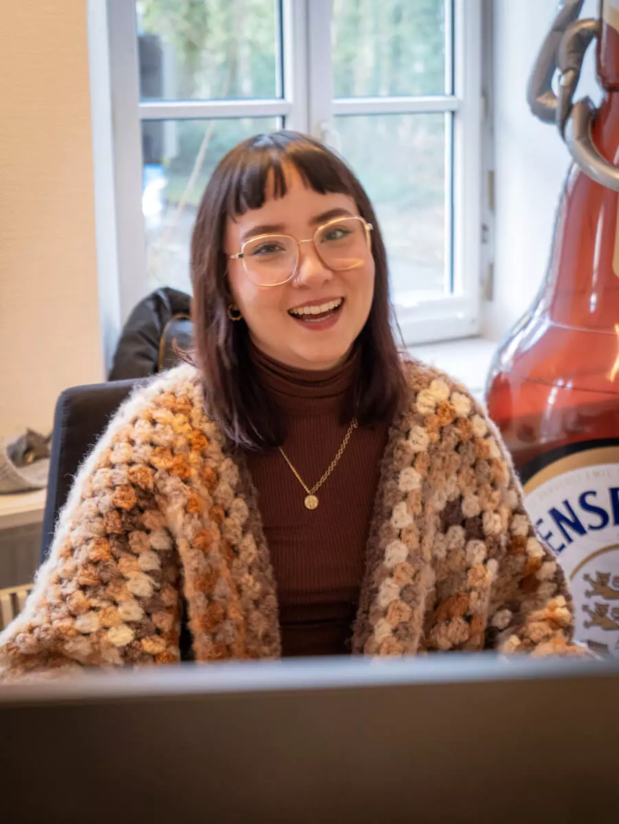 Eine Frau lächelt in ihrem Büro in die Kamera mit einer aufblasbaren Flensflasche im Hintergrund.